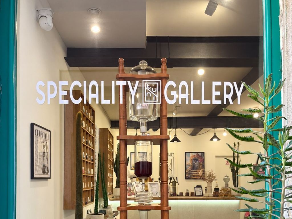 The window of Tunï Gallery with the lettering Speciality Gallery, behind it a wood and glass cold drip tower and shelves of porcelain cups