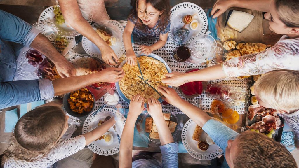 Familie mit Kindern von oben gesehen, alle greifen gemeinsam nach einer Pfanne in der Tischmitte, ringsum kleine Teller mit Brot, Käse, Schinken und Gebäck