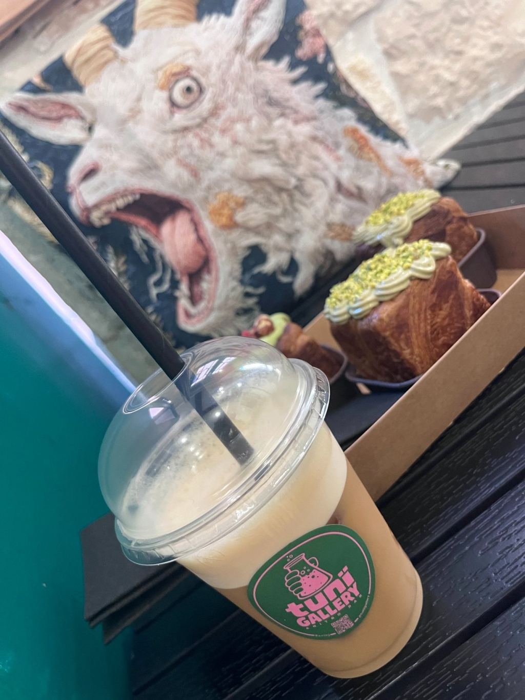 Cup of light coffee drink in front of a goat painting, next to cube croissants with pistachio topping