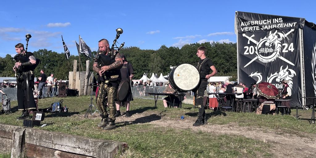 Zwei Dudelsackspieler und ein Trommler spielen auf einer Wiese, im Hintergrund Marktstände und ein MPS-Banner