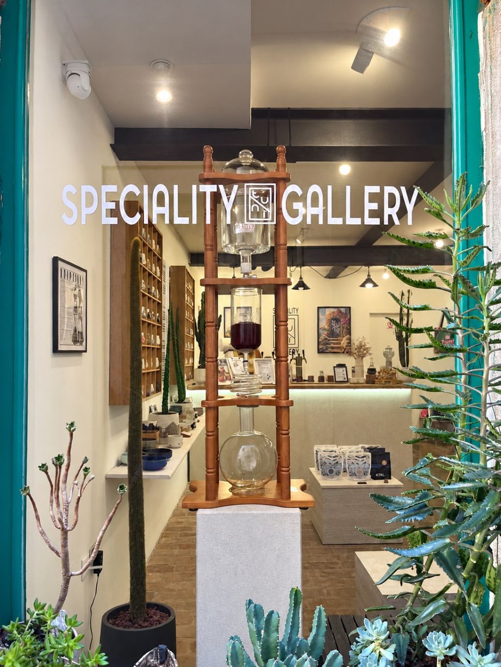 The window of Tunï Gallery with the lettering Speciality Gallery, behind it a wood and glass cold drip tower and shelves of porcelain cups