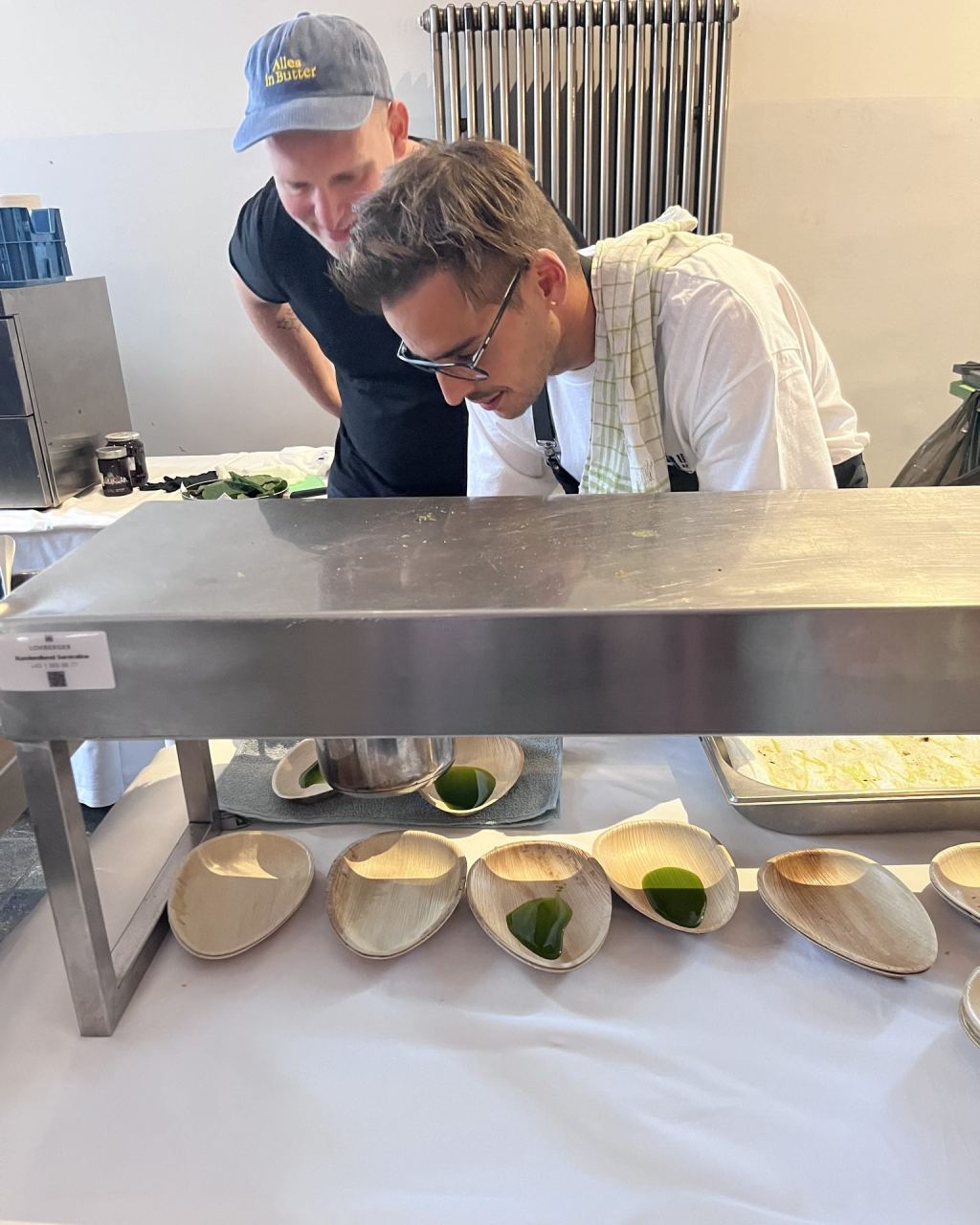 David Senfter und ein Mitarbeiter beugen sich über eine Wärmebrücke, darunter Palmblattschalen mit einem grünen Tropfen Sauce