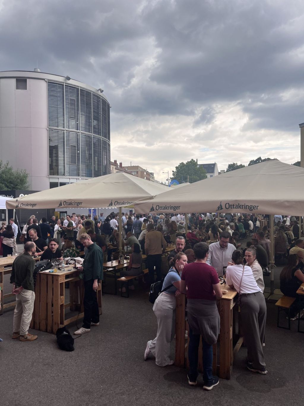 Viele Besucher an Stehtischen und Bänken unter großen Ottakringer-Schirmen im Außenbereich der Brauerei, dahinter ein runder Glasbau unter Wolken