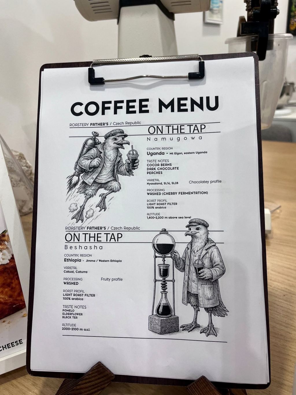 Coffee menu on a clipboard listing two cold drip coffees under the heading On the Tap, illustrated with birds