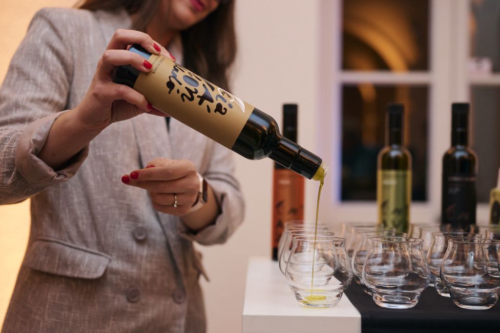 A woman pouring olive oil from a Chiavalon bottle into a tasting glass, with more glasses and bottles beside it