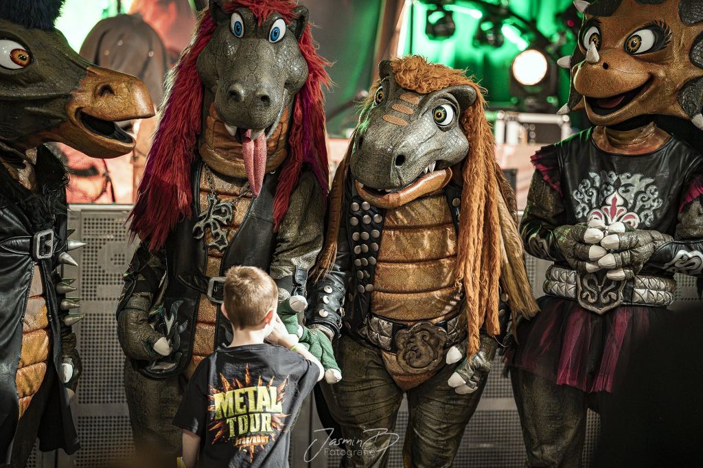 Ein kleiner Junge im Metal-Tour-Shirt steht beim Meet and Greet zwischen vier Heavysaurus-Dinos und schüttelt einem von ihnen die Hand