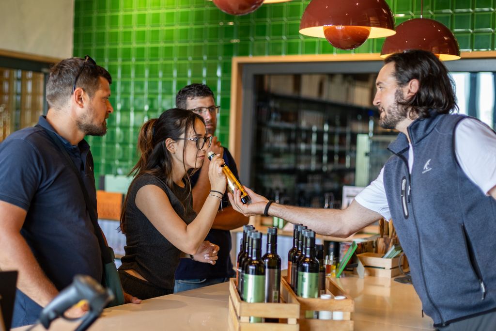 A Chiavalon employee hands a bottle of olive oil across the counter, a visitor smells it while two men look on, with bottles in wooden crates in front of them